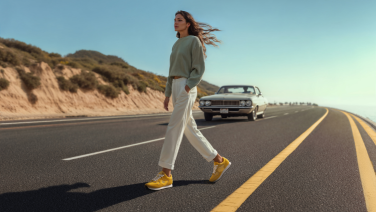 Woman in grey jumper and white trousers crossing the street wearing yellow trainers, vintage car in the background
