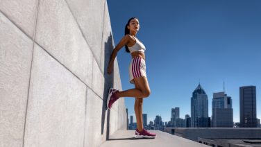 Woman in sportswear leaning against a concrete wall on a roof, wearing burgundy Wool Peak sneakers, with a city skyline in the background.