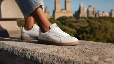 White sneakers on the feet of a person wearing dark trousers, sitting on a wall, with a city skyline in the background