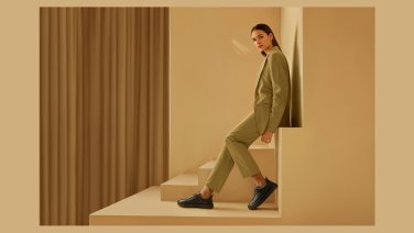 A young woman in an olive green suit sits on a beige platform in a minimalist room. She is wearing black shoes. The scene appears modern and elegant with clear geometric shapes.