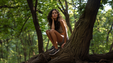 A young woman with long dark hair sits smiling on a thick tree branch in the forest, wearing dark trainers.