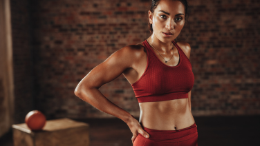 Sporty woman in red sportswear stands confidently with her hand on her hip in a gym in front of a brick wall.