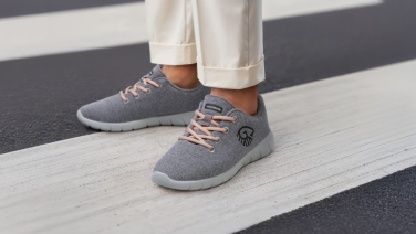 Feet in grey Giesswein trainers with orange laces stand on white zebra crossings. The person is wearing white trousers and the shoes feature the distinctive Giesswein logo.
