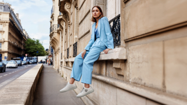 A woman in a light blue suit sits relaxed on a stone balustrade in front of classical architecture. She wears white trainers and smiles at the camera. An elegant city street can be seen in the background.