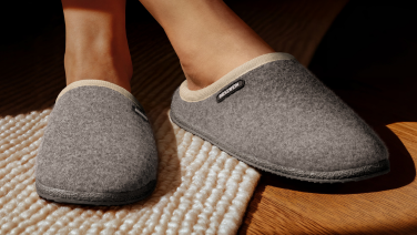 Close-up of feet in grey Giesswein slippers on a textured beige carpet. The slippers have a dark sole and feature the distinctive Giesswein logo.