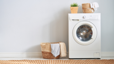 A white washing machine stands against a light-coloured wall in the laundry room. On top of the machine are a plant and some woven baskets. In front of it is a wicker basket containing woollen jumpers.