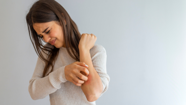 Woman with long brown hair wearing a beige knitted jumper scratches her forearm. She looks down at the itchy spot while scratching her arm with her other hand. Neutral grey background.
