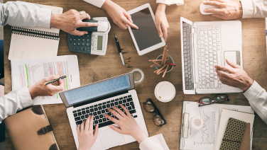 Several people work together at a table with laptops, tablets, calculators and documents - a busy team atmosphere.