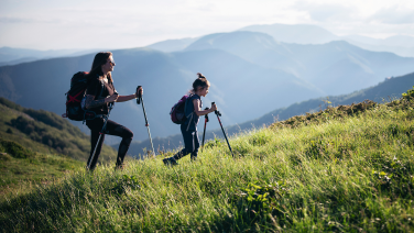 Two hikers with backpacks and walking sticks climb a grassy mountainside. In the background, a vast mountain landscape stretches out with several mountain ranges in shades of blue under a cloudy sky.