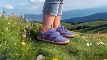 Purple children's shoes with Velcro fastenings on a green meadow with colourful wildflowers. In the background, a hilly landscape stretches out with rolling green hills under a blue sky.
