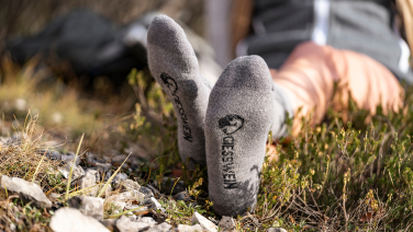 A person is lying on their back in the grass, feet stretched out in front of them, wearing grey hiking socks with a black logo and the word “GIESSWEIN”. Hiking clothes and nature can be seen blurred in the background.