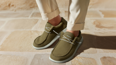 Close-up of feet in white trousers and olive green shoes with white laces and light-coloured soles. The person is standing on a light-coloured stone floor.