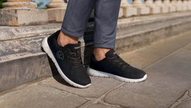 Close-up of black Giesswein Merino trainers on the feet of a person wearing grey jeans, standing on a stone wall in an urban setting.
