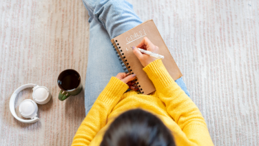 Top view of person in yellow jumper with notebook on carpet, coffee and coffee cup next to it