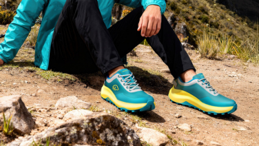 Person sitting on rocks in the mountains wearing turquoise and yellow Giesswein trainers, black trousers and a turquoise jacket.