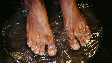 The bare feet of a person standing in clear, splashing water. Water droplets and splashes are frozen in motion, emphasising the refreshing and natural atmosphere.