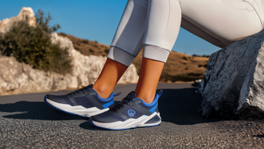 Close-up of a Giesswein sneaker in dark blue with blue accents on a woman's ankle. The person is wearing light grey leggings and is standing on asphalt in front of a rocky background with a blue sky.