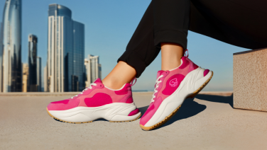 A close-up of pink Giesswein trainers with white chunky soles on a person’s feet. Skyscrapers and a blue sky can be seen in the background.
