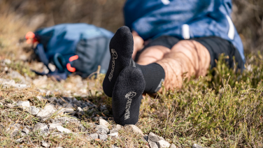 A person is lying relaxed on a rocky surface in the countryside. In the foreground, you can see their feet wearing black Giesswein merino socks.