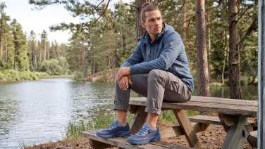 A man in a blue jacket is sitting on a wooden bench by the lake; he is wearing blue merino wool walking boots