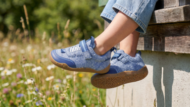 Close-up of light blue Giesswein children's trainers with hook-and-loop fastener and honey-coloured sole, worn with jeans in front of a blooming summer meadow.