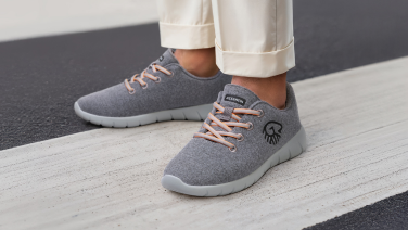 Close-up of a person's feet in cream-coloured, rolled-up trousers. They are wearing grey merino wool trainers with apricot-coloured laces and a light grey sole. The person is standing on a zebra crossing.