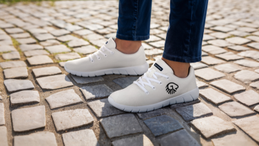 Close-up of white Giesswein merino trainers with a black brand logo on the feet of a person in jeans, standing on cobblestones.