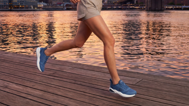 A person wearing beige sportswear is running along a wooden jetty by the water at sunset; the outline of a town can be seen in the background. They are wearing blue, sporty running shoes.