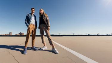A couple on a sunny roof terrace with a panoramic view of the city. He is wearing a denim jacket, beige shorts and blue merino wool trainers; she is wearing a dark green jacket, beige trousers and grey trainers.