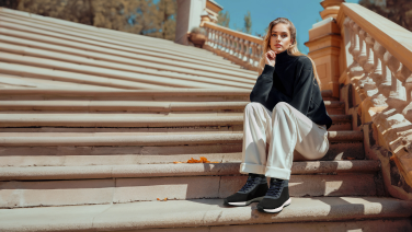 Woman in black jumper and white wide-leg trousers sitting on sandstone-coloured steps with classic banister, wearing black Giesswein trainers.