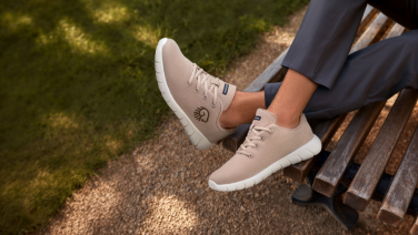 Close-up of a beige Giesswein Merino trainer with a white sole and embossed logo on the feet of a person in grey trousers on a park bench.