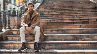 Man in beige jacket and light-coloured trousers sitting on stone outdoor steps wearing black Giesswein winter trainers, autumnal atmosphere