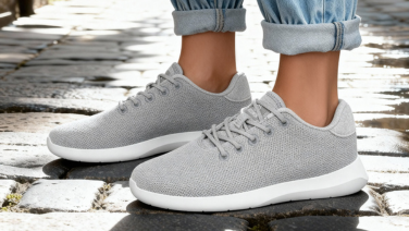 Close-up of light grey Giesswein Merino trainers with white soles on cobblestones, worn with rolled-up jeans in sunlight.