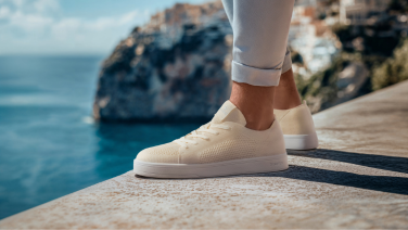 Close-up of a cream-coloured Giesswein Merino trainer with a white flat sole on a stone wall against a Mediterranean sea backdrop.