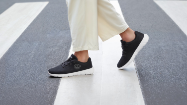 Close-up of black Giesswein merino trainers with a white sole on the feet of a person in white wide-leg trousers on a zebra crossing.