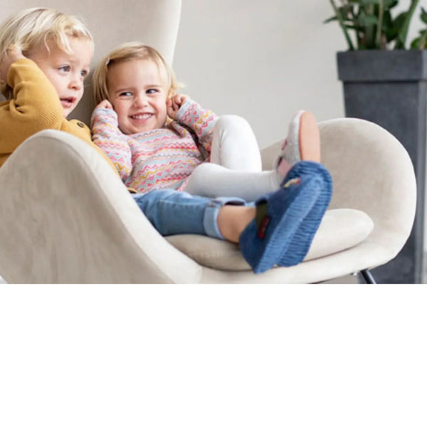 Two toddlers are sitting together in a beige armchair. The child in front is wearing blue knitted slippers and smiling happily.
